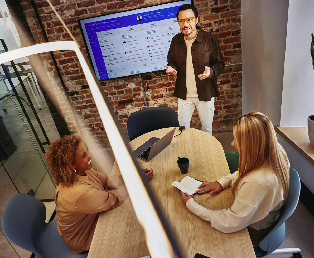 an image of three people in a meeting room with a huge screen showing data and one of them standing and discussing 