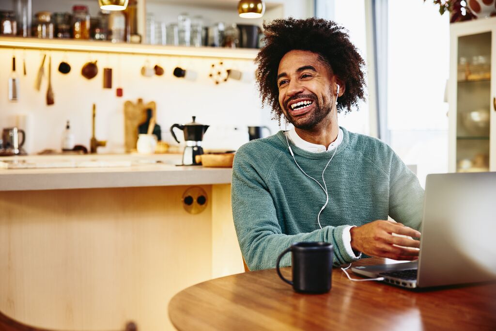 Smiling man sitting at his dining table with a drink and his laptop. Looking away. Kitchen in the background.