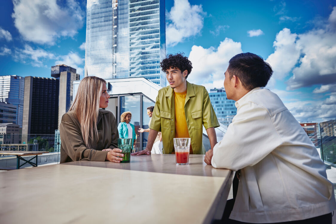 an image of three people, a male in yellow shirt standing, female wearing a brown long sleeved top and another male in a white jacket sitting in a chair outside on a balcony at a table, talking