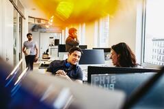 a man with headphones placed at his neck talking to a woman in the office  while there are people behind them