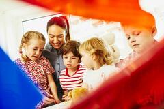 A woman with kids and toys, a teddy bear and colored pencils. Window in the background.