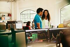 A man and woman chatting in the office with computer screens showing graphs in front of them