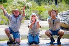 an image of three smiling people wearing collared shirts and hats trying to catch water in their palms while crouching and kneeling down