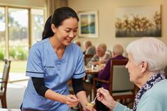 a smiling aged care professional wearing scrubs in an aged care facility serving a resident her lunch