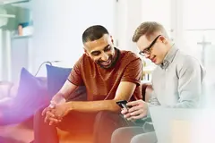 Two people sitting on a couch smiling while looking at a phone.

