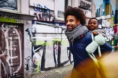 Smiling male with kid on his back walking through a city.
