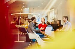 Group of colleagues sitting at a table in a meeting with a laptop, talking and smiling. Office and other people in the background.