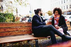 man and woman sitting on a bench, having coffee and laughing during conversation. 
