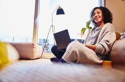 Smiling woman leaning against a couch, sitting on the floor, with laptop on her lap