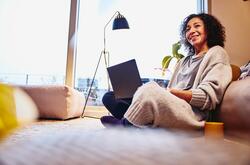 Smiling woman leaning against a couch, sitting on the floor, with laptop on her lap.