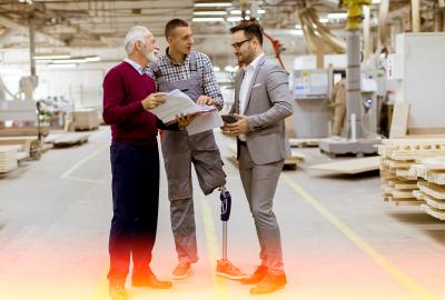 A man with a prosthetic leg stands in a factory talking to two colleagues about information in a folder.