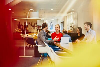 Group of colleagues sitting at a table in a meeting with a laptop, talking and smiling. Office and other people in the background.