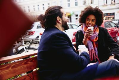 Smiling man and woman with drinks sitting down on bench outside. Having a conversation.