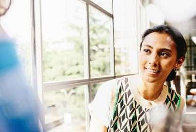 a woman in a colourful top seated by the window while talking to her colleague