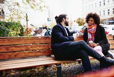 man and woman sitting on a bench, having coffee and laughing during conversation. 