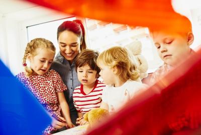 A woman with kids and toys, a teddy bear and colored pencils. Window in the background.