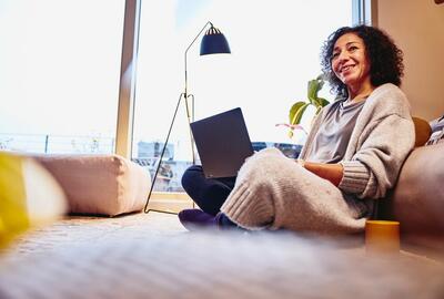 Smiling woman leaning against a couch, sitting on the floor, with laptop on her lap