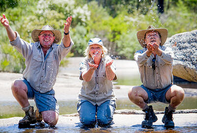 an image of three smiling people wearing collared shirts and hats trying to catch water in their palms while crouching and kneeling down