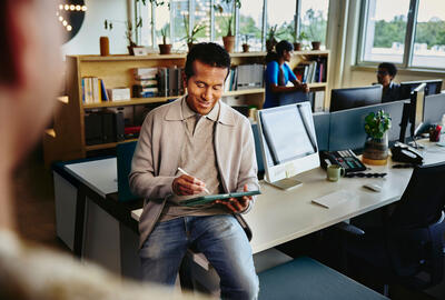 an image of a man writing on his tablet while seated on a desk smiling