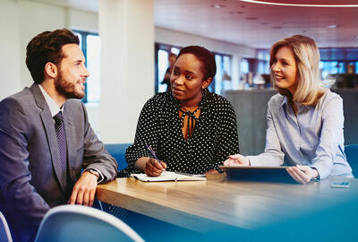 an image of three colleagues chatting while sitting down and one is taking notes