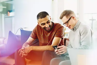 Two people sitting on a couch smiling while looking at a phone.
