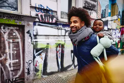 Smiling male with kid on his back walking through a city.
