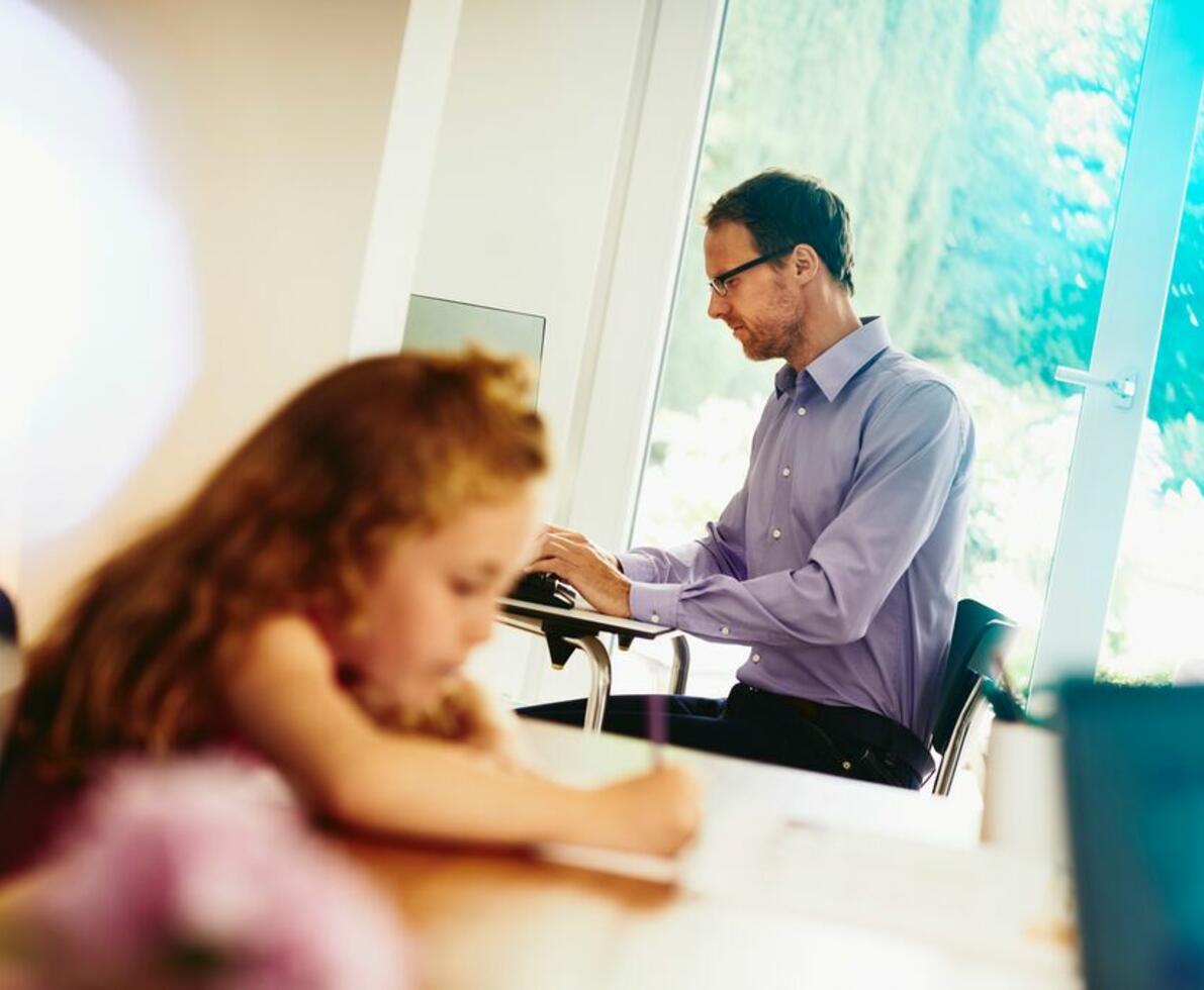 an image of a man working at home and using a laptop while his daughter is writing something on a desk