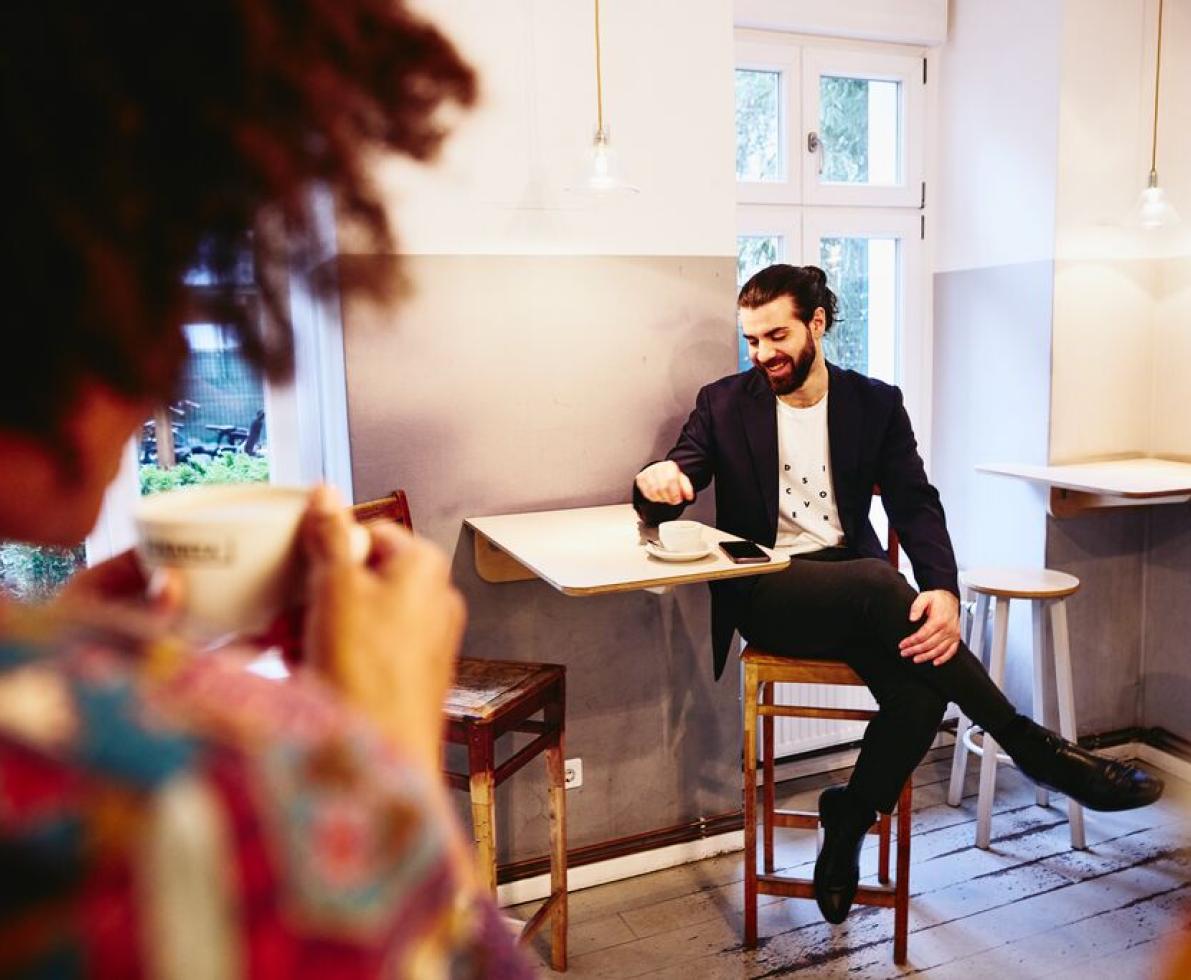 Man sitting at a table with coffee, looking down while having a conversation with someone.