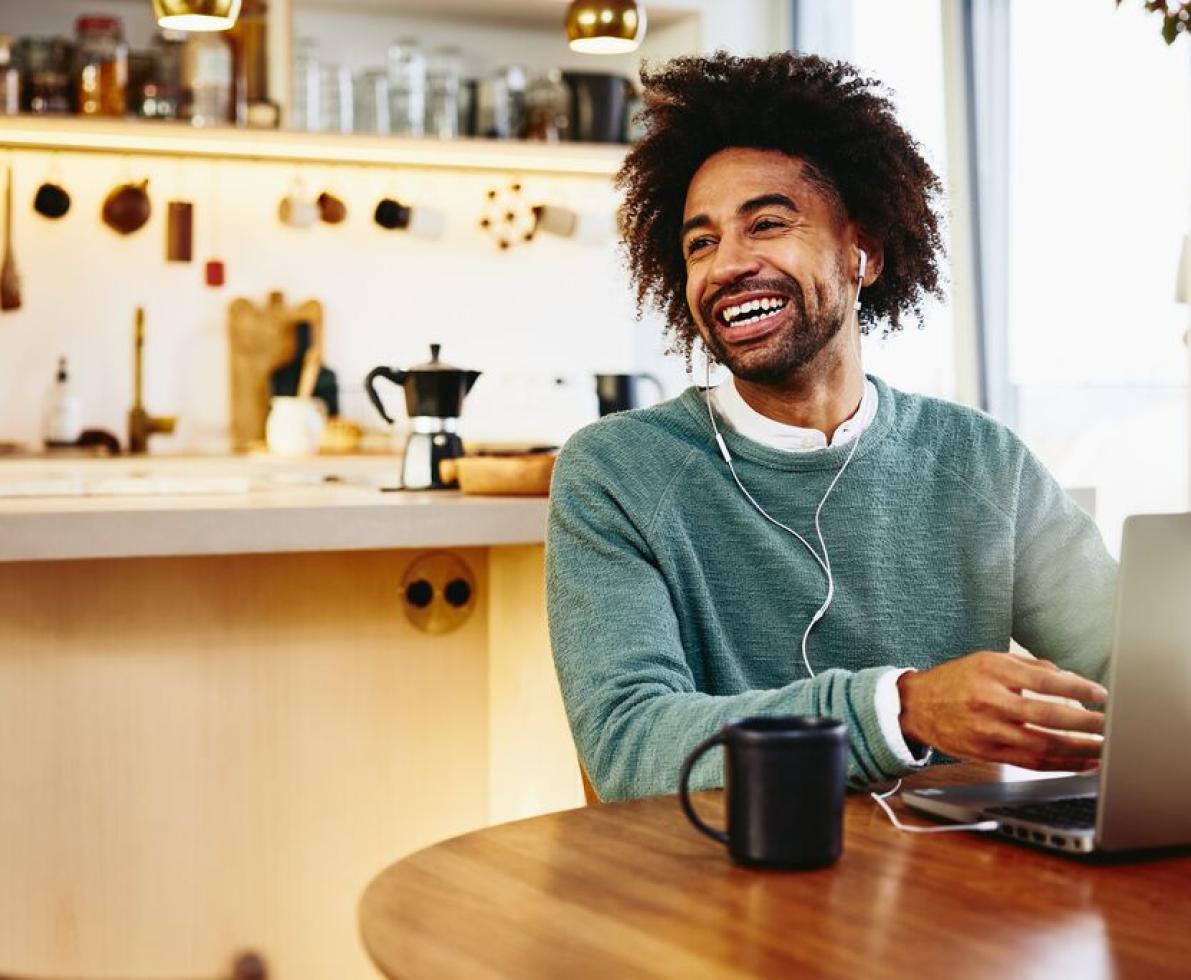 Smiling man sitting at his dining table with a drink and his laptop. Looking away. Kitchen in the background.