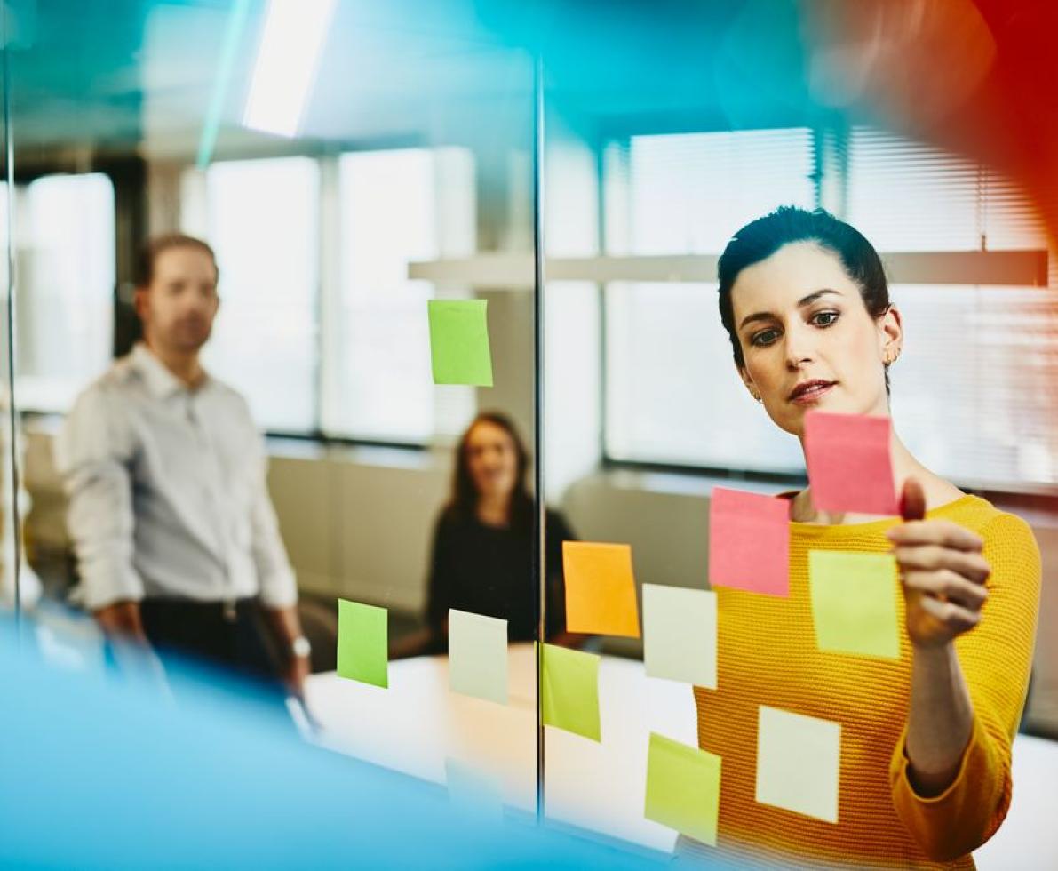 Two business women and a man in an office putting sticky notes on a window.