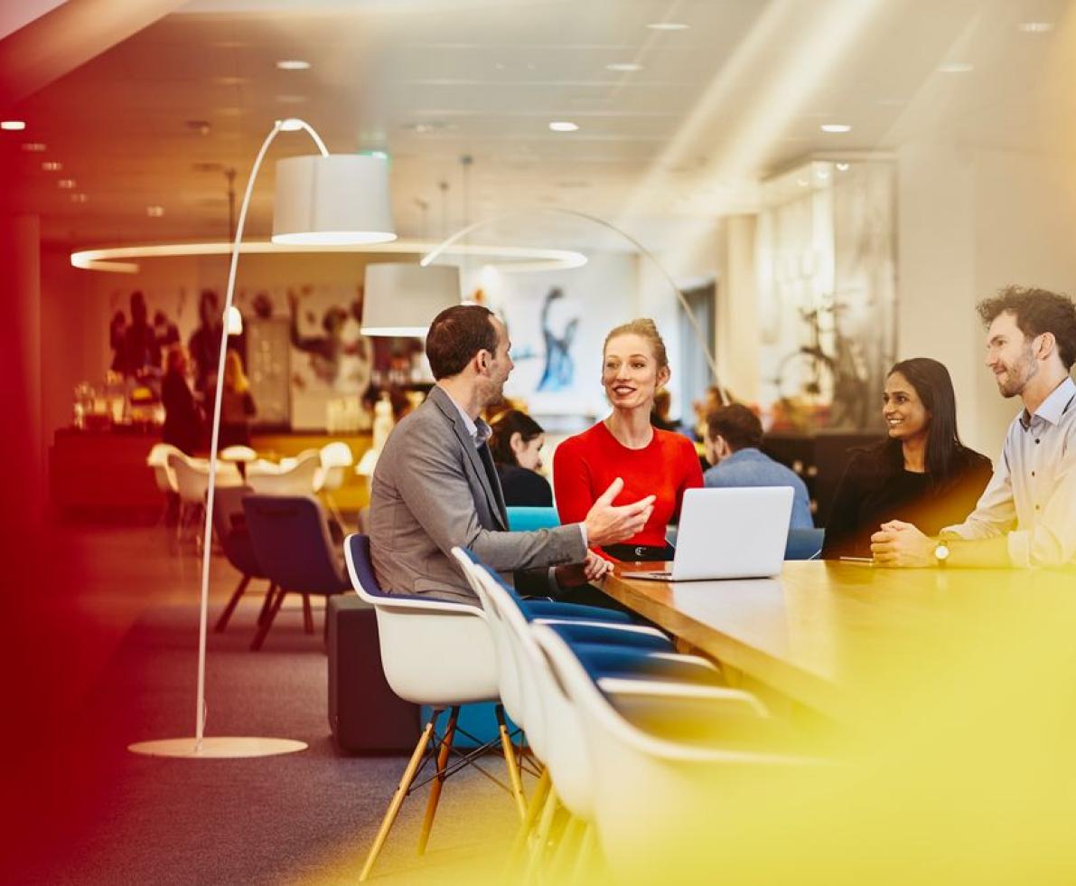 Group of colleagues sitting at a table in a meeting with a laptop, talking and smiling. Office and other people in the background.