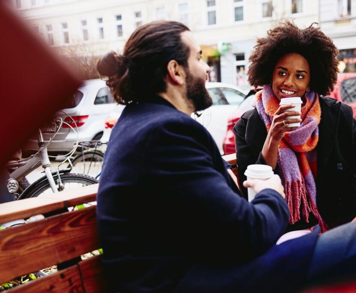 Smiling man and woman with drinks sitting down on bench outside. Having a conversation.