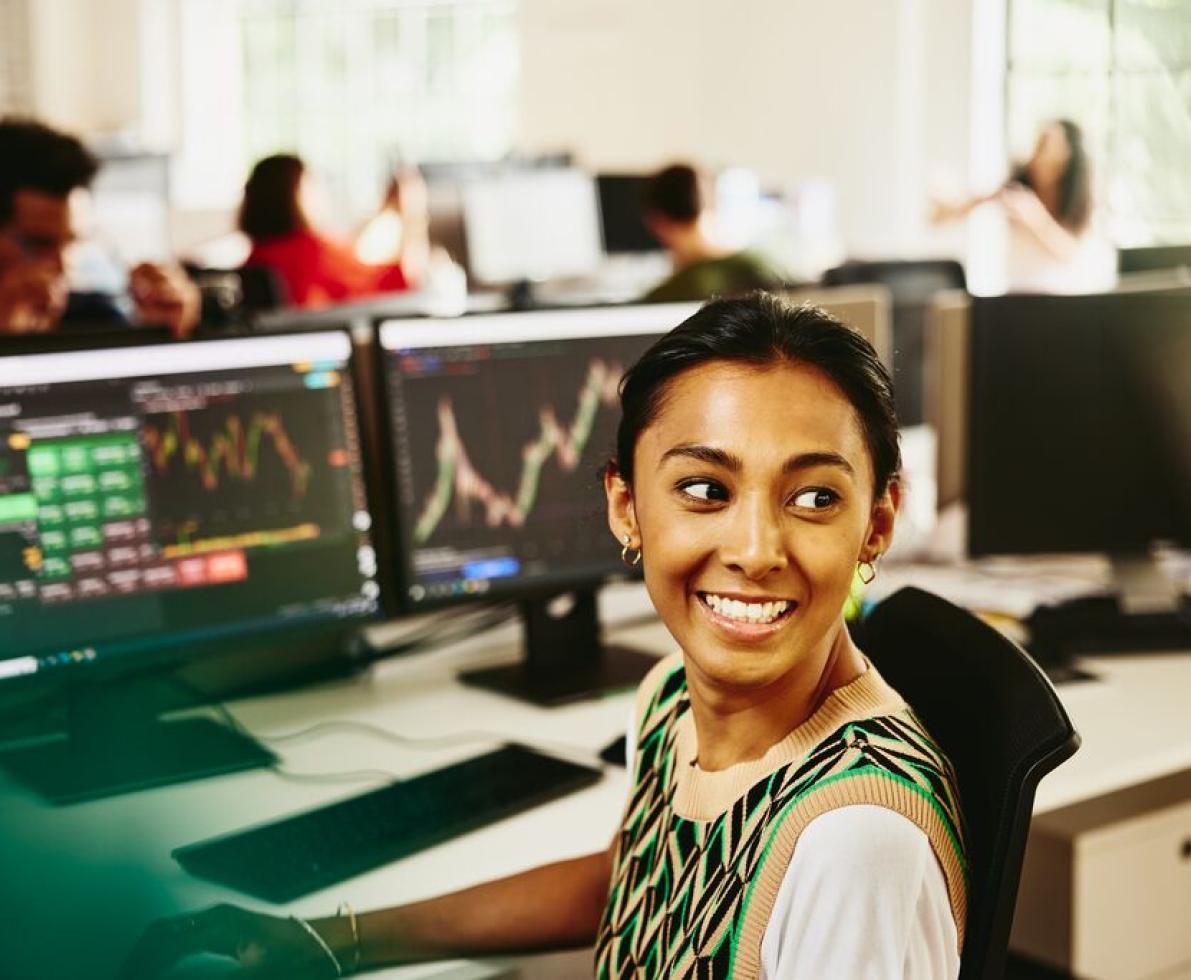 Smiling woman looking away from computer screens displaying financial information.