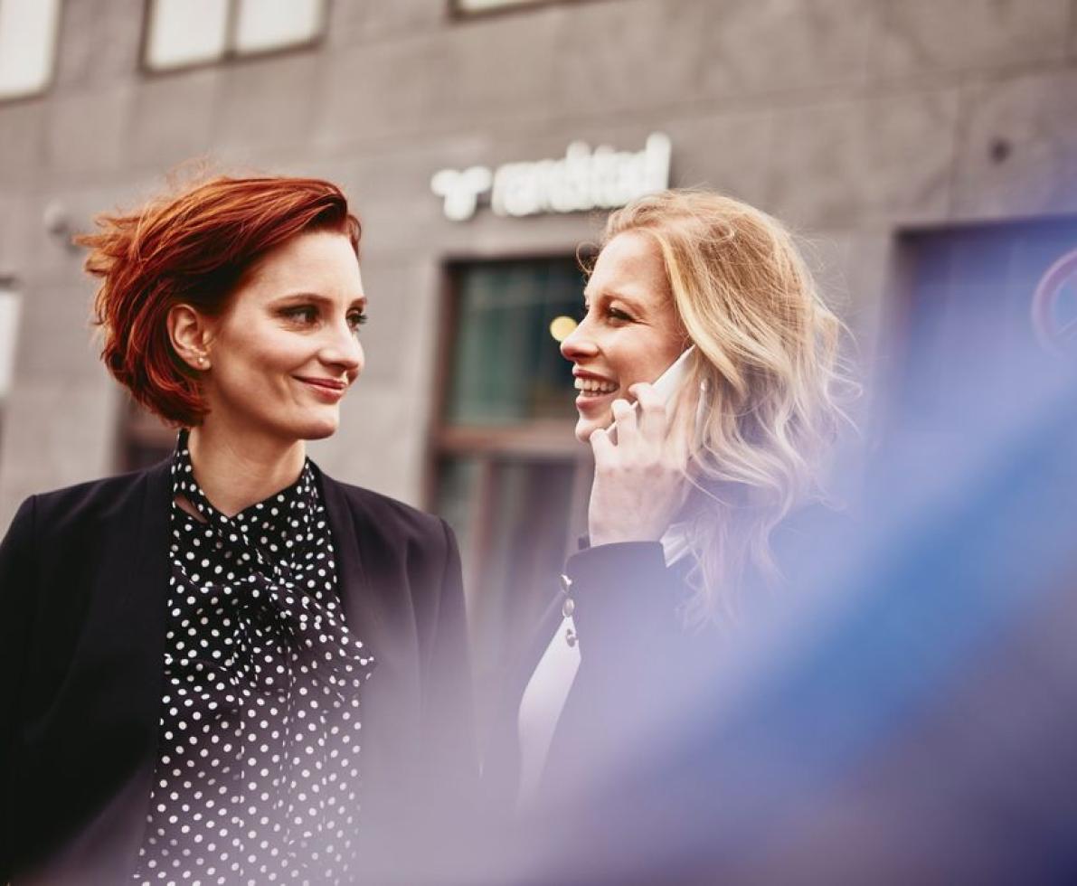 Two smiling woman, talking and walking around the city. One woman is on the phone.