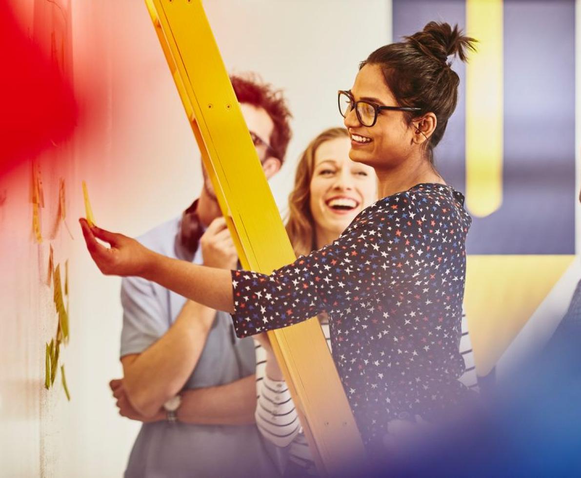 Woman standing on a yellow ladder in an office, sticking a post it on a whiteboard. Group of colleagues around her. Smiling Woman standing on a yellow ladder in an office, sticking a post it on a whiteboard. Group of colleagues around her. Smiling