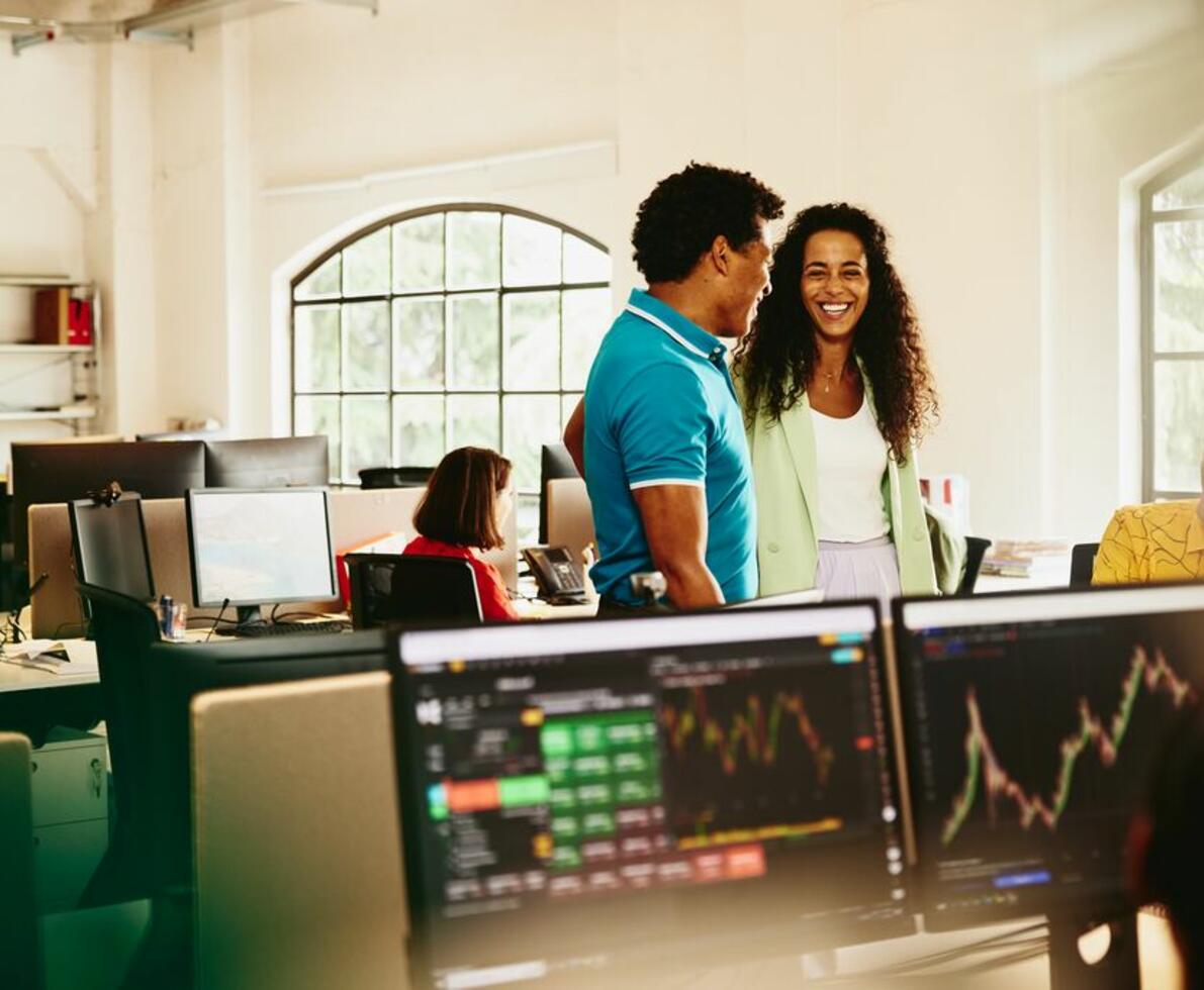 A man and woman chatting in the office with computer screens showing graphs in front of them