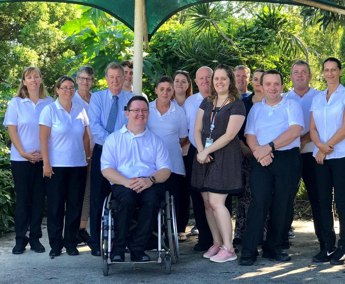 the gold coast health team of 13 people standing in front of greenery, one is in a wheelchair