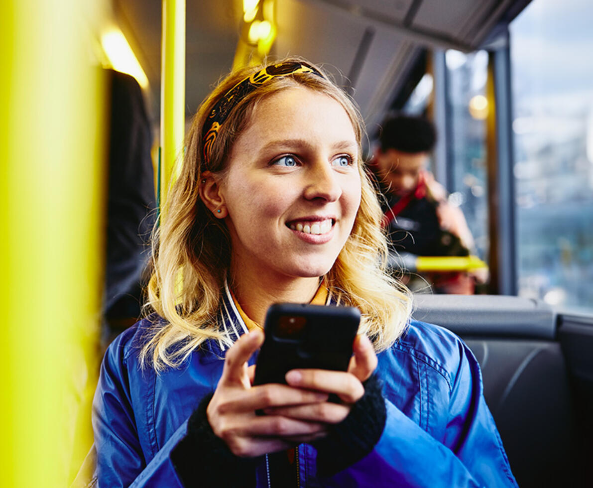 an image of a woman on a bus, smiling while looking out the window and holding her phone