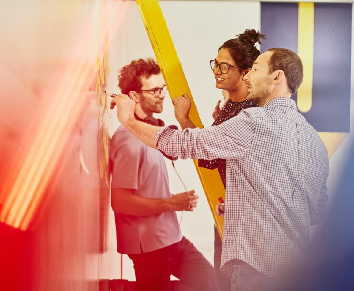 Woman standing on a yellow ladder in an office. Man writing on a whiteboard