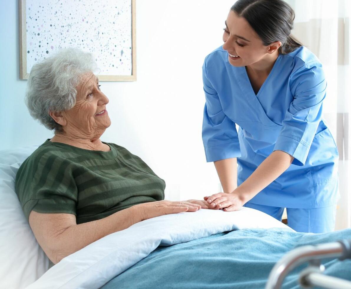 Carer talking to her patient who is sitting up in bed. Both are smiling and the carer has her hand on the patient's hand.
