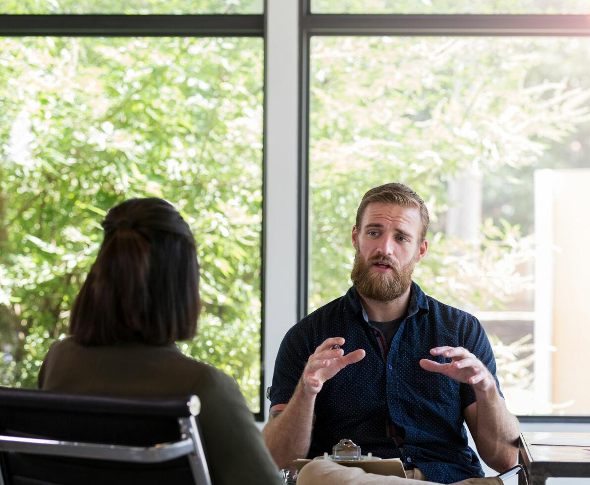 Therapist in an office setting talking to his patient. Both are sitting next to a window. 