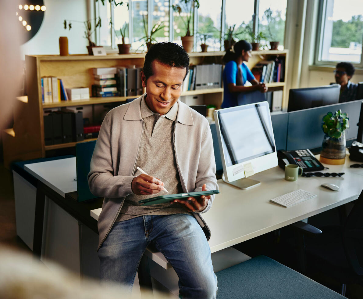 an image of a man writing on his tablet while seated on a desk smiling