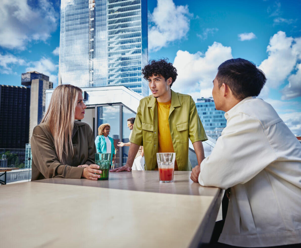 an image of three people, a male in yellow shirt standing, female wearing a brown long sleeved top and another male in a white jacket sitting in a chair outside on a balcony at a table, talking