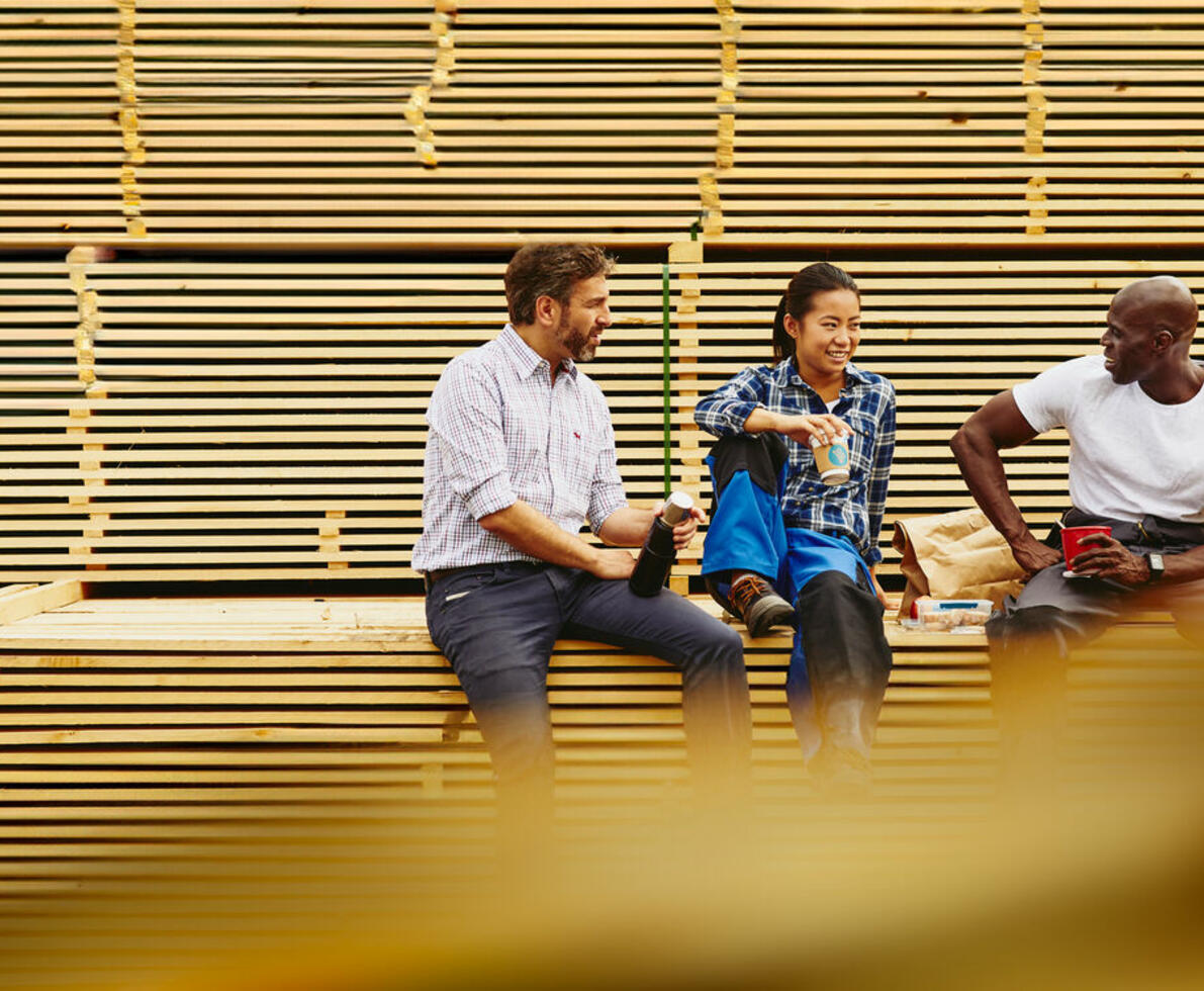 an image of three colleagues chatting during a lunch break while sitting on a pile of wood.
