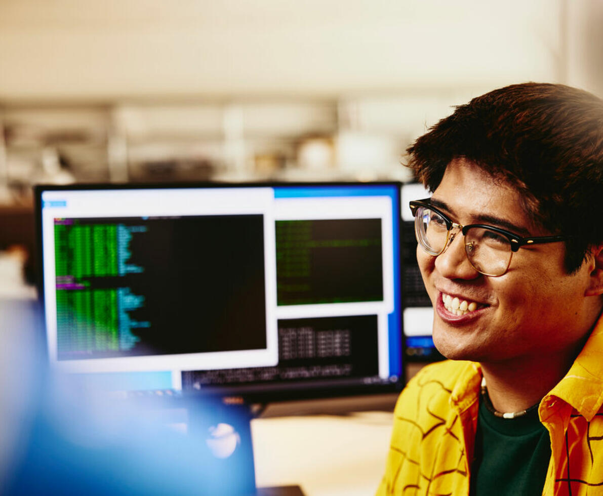 a man wearing glasses sitting behind his desk, computer screens displaying programming code.