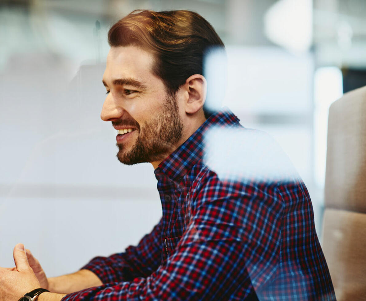 a bearded man wearing a chequered collared shirt smiling with his hands clasped in front of him