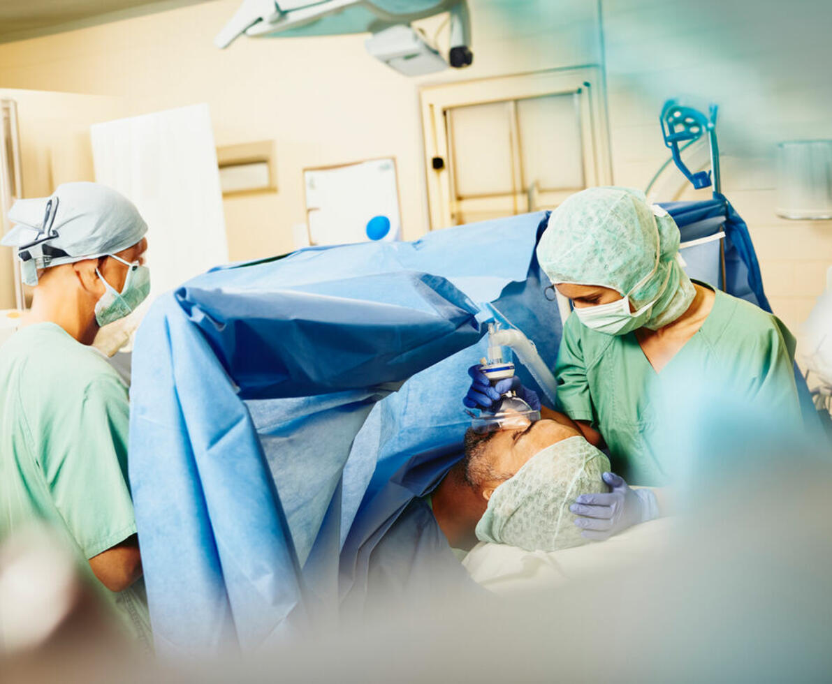 Two nurses in an operating room. There is a patient on the operating table. One of the nurses is holding an oxygen mask over the patient's mouth