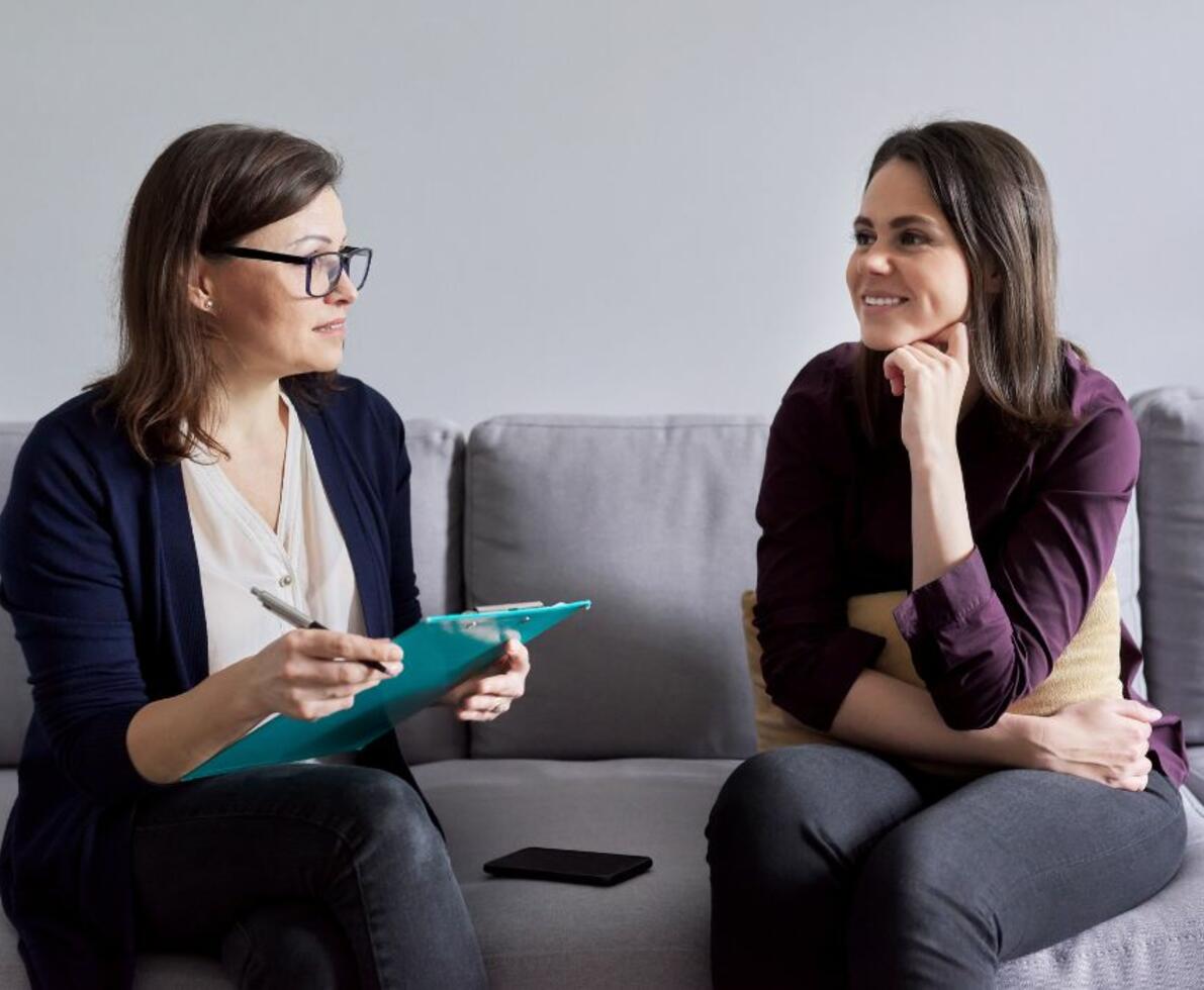 image of a social worker having a consultation with her patient. They are sitting on a grey couch. The patient is smiling.
