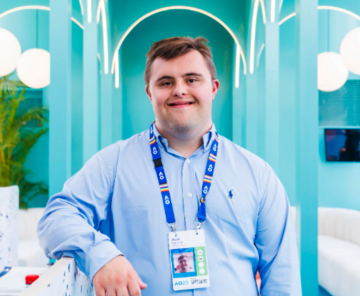 man with short brown hair, wearing a name tag wearing a light blue shirt and black pants standing next to a reception desk