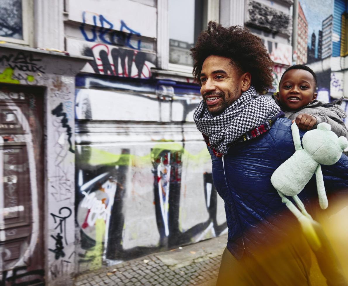 Smiling man with kid on his back walking through a city.